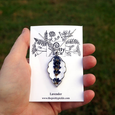 Lavender Sprig Necklace held in hand in a grass field setting, showcasing pendant design, craftsmanship, and natural lifestyle aesthetic