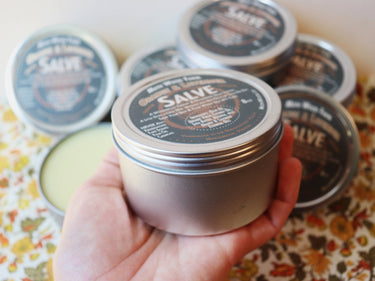 Orange & Lavender Salve tin in a bigger size held in hand with full-size tins in the background, highlighting product variety and packaging design