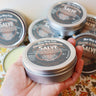 Orange & Lavender Salve tin held in hand with multiple tins displayed in the background showcasing product packaging and branding