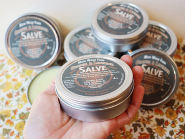Orange & Lavender Salve tin held in hand with multiple tins displayed in the background showcasing product packaging and branding