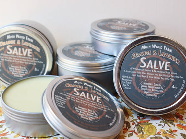 Orange & Lavender Salve tin open showing smooth cream inside, with another tin beside it displaying the product label and packaging design